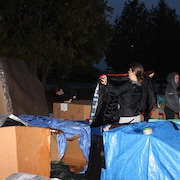 Une personne tient un sac de couchage dans un campement de boîtes de carton.