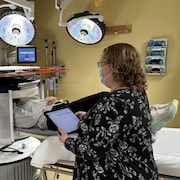 Une femme portant un masque médical tient une tablette numérique dans ses mains pendant qu'une patiente est couchée sur le dos lors d'un examen par imagerie par résonance magnétique.