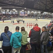 Un aréna rempli de spctateurs assis et debours devant la glace où sont alignés les joueurs des deux équipes de hockey pendant que des dignitaires sont sur le tapis rouge avant le match. 
