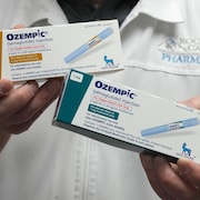 Un homme vêtu d'un sarrau de pharmacien tient dans ses mains deux boîtes d'Ozempic, qui affichent des informations sur le médicament et la photo d'un injecteur.