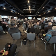 Une foule assise dans une grande salle lors d'une assemblée générale annuelle. 