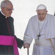 Le pape muni d'une canne est aidé par un cardinal.