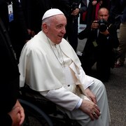 Le pape est assis sur une chaise roulante et est entouré de photographes.