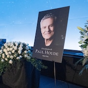 Une affiche qui montre Paul Houde souriant est entourée de bouquets de roses lors de la chapelle ardente.