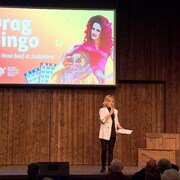 Monique Poirier sur scène devant un public au Pays de la Sagouine. Derrière elle, un immense écran qui porte la mention drag bingo avec Rose Beef et Joséphine.