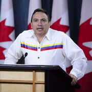 Perry Bellegarde parle sur une tribune devant une série de drapeaux canadiens.