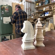 Deux figurines de bois en forme de pion d'échec sont posées sur une table et Pierre-Luc Gauthier travaille sur une machine en arrière.