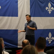 Debout devant un immense drapeau du Québec, Pierre Poilievre s'exprime au micro devant des partisans. 
