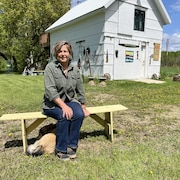 Une personne assise sur un banc, un chien à ses pieds et une grange à l'arrière. 