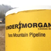 Logo de l'entreprise Kinder Morgan en premier plan, en bordure d'une route. L'entreprise Kinder Morgan affirme que l'attente des permis d'expansion du pipeline Trans Mountain, en Colombie-Britannique, a des répercussions coûteuses pour le projet. 