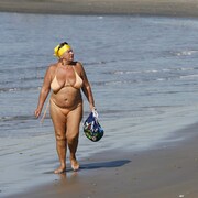 Une femme en bikini couleur chair, un foulard jaune sur la tête, marche sur le sable près de l'eau, un sac à la main.