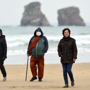 Trois personnes, dont deux masquées, marchent sur une plage.