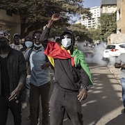 Des manifestants masqués déambulent dans une rue du quartier du Plateau au Sénégal. De la fumée émerge d'une voiture garée en bordure.