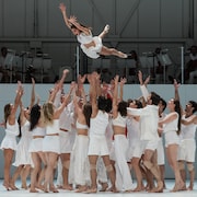 Sur une scène, une danseuse tombe des airs dans les bras d'une vingtaine d'autres danseurs.