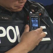Zoom sur la caméra, plus petite qu'un téléphone cellulaire, sur le gilet d'un policier.