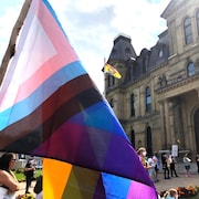 Des gens sont regroupés devant l'édifice de l'Assemblée législative du Nouveau-Brunswick. Un grand drapeau transgenre flotte au vent.