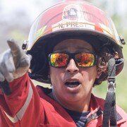 Une pompière casquée, en uniforme, pointe devant elle. Dans ses lunettes fumées, on voit d'autres pompiers devant un feu de forêt.