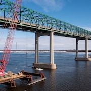 Un pont enjambe le fleuve près d'une grue rouge installée sur une structure qui surplombe l'eau.