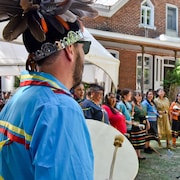 Un homme en costume traditionnel abénaquis chante avec un tambour à la main. 