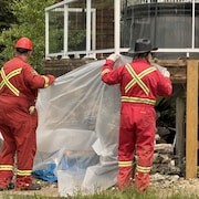 Des pompiers mettent du plastique autour de bois sec entreposé sous une maison de Candle Lake, en Saskatchewan, dans le cadre du programme FireSmart pour protéger les habitations d'un feu de forêt. (10 juin 2025)