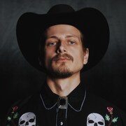 Le chanteur pose avec un chapeau de cowboys et une chemise avec des têtes de mort et des fleurs.