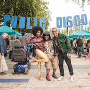 Trois personnes posent sur une place publique, de manière amusante, devant une banderonle sur laquelle est écrit Public Disco.
