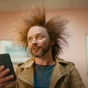 Scène de la publicité. Les cheveux du personnage se soulèvent dans les airs après qu'il a vu sur son téléphone intelligent que l'Ontario investit dans la production de batteries pour véhicules électriques.