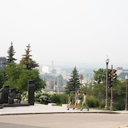 Vue lointaine de Québec sous un ciel brumeux et voilé. Trois personnes marchent sur un trottoir. Un feu de signalisation rouge est visible à droite, avec un panneau vert indiquant un virage à droite autorisé. Des monuments et des arbres sont visibles sur la gauche, avec des bâtiments en arrière-plan. La visibilité est réduite en raison de la fumée.