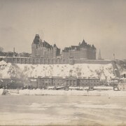 Québec vue de Lévis, en hiver. Sa tour centrale n'est pas encore construite. Le cap est couvert de neige. On distingue des chevaux attelés à des banneaux à neige près de l'imposant marché Champlain situé près du fleuve.
