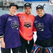 Trois joueurs de hockey queer en uniforme sur la glace sourient à la caméra.