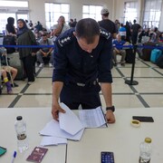 Un agent consulte une liste à une table, devant des gens qui attendent dans une salle. 