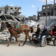 Deux hommes, une femme voilée et une enfant assis sur un chariot tiré par un cheval émacié circulent dans une rue devant des immeubles détruits.
