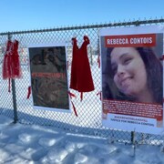 Des pancartes, dont l’une affiche la photo de Rebecca Contois, et des robes rouges sont accrochées sur une clôture grillagée l’hiver.