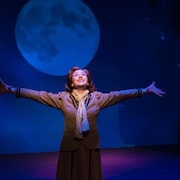 Une femme est au milieu de la scène du théâtre, bras ouverts, et regarde la lune qui se trouve au-dessus d'elle.