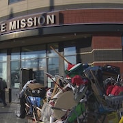 Des personnes sont debout devant un bâtiment qui porte l'écriteau Hope Mission sur la devanture. Un chariot de supermarché avec des affaires entassées est stationné devant l'immeub.