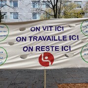 Une banderole sur laquelle est écrite en plusieurs langues la phrase « On vit ici, on travaille ici, on reste ici ».