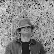 L'homme portant un chapeau se tient devant des arbustes.