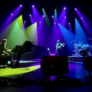 Une scène illuminée où se produisent des artistes musicaux, tels une pianiste, un guitariste et un batteur.
