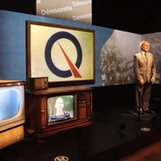 Une statue de René Lévesque en exposition avec deux vieux téléviseurs.