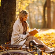 Une femme lit un livre adossée sur un arbre pendant l'automne, avec une bicyclette couchée sur le sol près d'elle. 