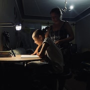 Un enfant étudie alors que sa mère est penchée derrière lui.