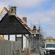 Trois personnes sur des échafauds travaillent à réparer le toit et la cheminée de la maison Rodrigue.