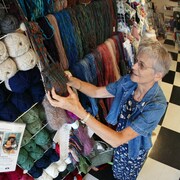 Une dame âgée tient un sac tricoté dans ses mains, dans un magasin où sont exposées des pelotes de laine de toutes les couleurs. 