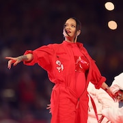 Une femme au ventre arrondi porte une combinaison rouge, entourée de danseurs habillés de blanc, lors d'un spectacle.