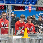 Des jeunes garçons avec le même chandail d'équipe contrôlent leur robot lors d'une compétition. Des gens autour regardent . 