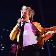 Un homme vêtu d'un veston orange chante dans un micro sur scène.