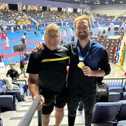 Roger et Alexandre St-Arneault, qui montre sa médaille d'or, posent pour la photo dans un aréna. Une compétition de karaté se déroule à l'arrière-plan.