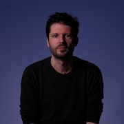 Le journaliste Romain Schué est en studio, assis devant un mur, et il regarde l'objectif de la caméra.