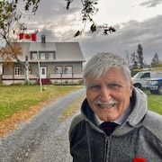 Roméo Dallaire sourit tandis qu'on voit une maison à l'arrière avec, juste devant, un drapeau du Canada flottant au vent.