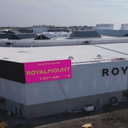 Un immense édifice commercial sur lequel est apposé une banderole avec la mention « Vous êtes invités Royalmount 5 sept 2024 ».